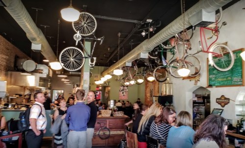 A bustling café with bicycles hanging from the ceiling and patrons enjoying their time at tables and the counter.