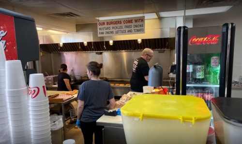 A busy burger joint kitchen with staff cooking, a soda machine, and a sign listing burger toppings.
