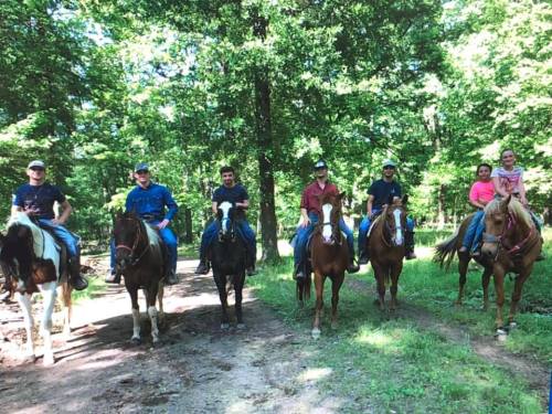 A group of six people on horseback, riding through a wooded area on a sunny day.