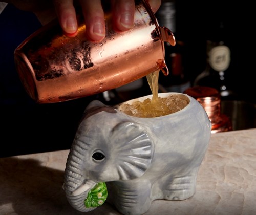 A cocktail being poured from a copper shaker into a ceramic elephant-shaped mug filled with ice.