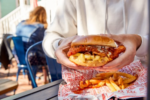 A person holds a spicy chicken sandwich with pineapple and fries on a table in a casual dining setting.