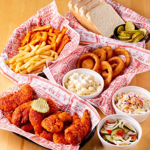 A spread of fried chicken, fries, onion rings, macaroni salad, coleslaw, pickles, and slices of bread in red-lined baskets.