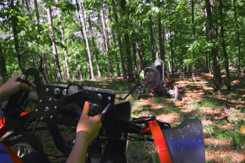 A person driving an ATV through a lush green forest, with another vehicle visible in the background.