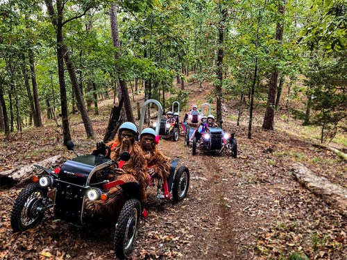 A group of people in costumes driving small off-road vehicles through a wooded trail.