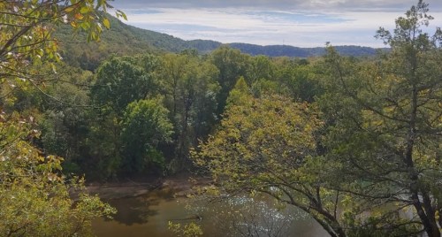 A serene landscape featuring a river surrounded by lush green trees and rolling hills under a cloudy sky.