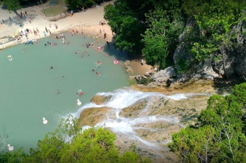 Aerial view of a waterfall cascading into a swimming area with people enjoying the water and surrounding greenery.