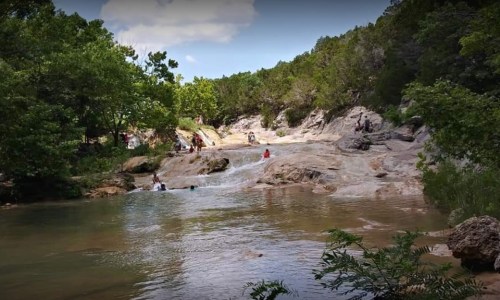 A scenic view of a rocky stream with people enjoying the water and lush greenery surrounding the area.