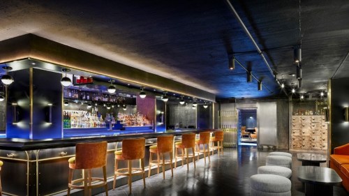 A modern bar interior featuring a long counter, stylish seating, and illuminated shelves filled with bottles.