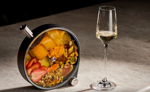 A round black container filled with colorful fruits and nuts, next to a glass of sparkling wine on a marble surface.