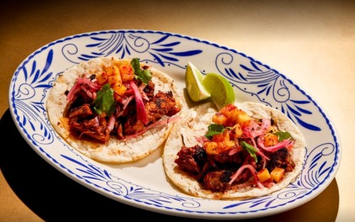 Two tacos on a decorative plate, filled with meat, pineapple, pickled onions, and garnished with lime wedges.