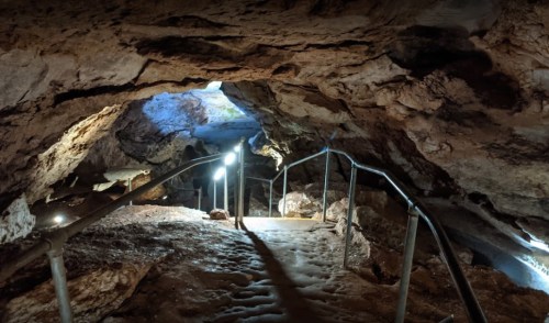 A dimly lit cave entrance with a metal railing leading down a rocky path.