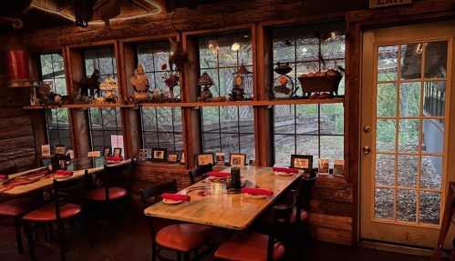 Cozy restaurant interior with wooden decor, tables set for dining, and large windows overlooking a natural view.