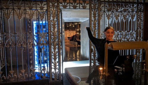A smiling receptionist stands at a stylish entrance with ornate metalwork, welcoming guests into a modern space.