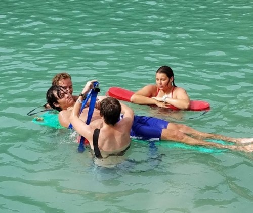 Two people assist a third on a float in the water, while another person watches nearby.
