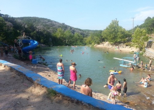 A sunny swimming area with people enjoying the water, surrounded by trees and a sandy shore.