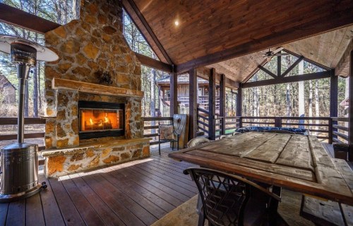 Cozy outdoor space with a stone fireplace, wooden table, and views of trees through a covered porch.