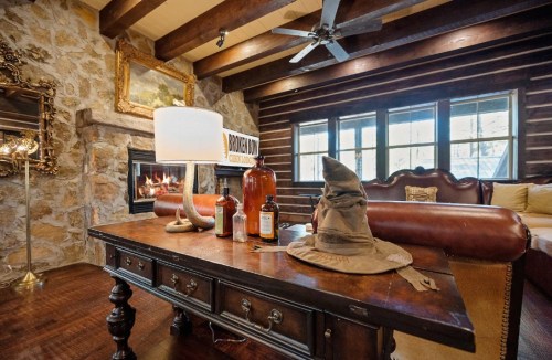Cozy living room with a stone fireplace, wooden beams, a table with a lamp, bottles, and a wizard hat.