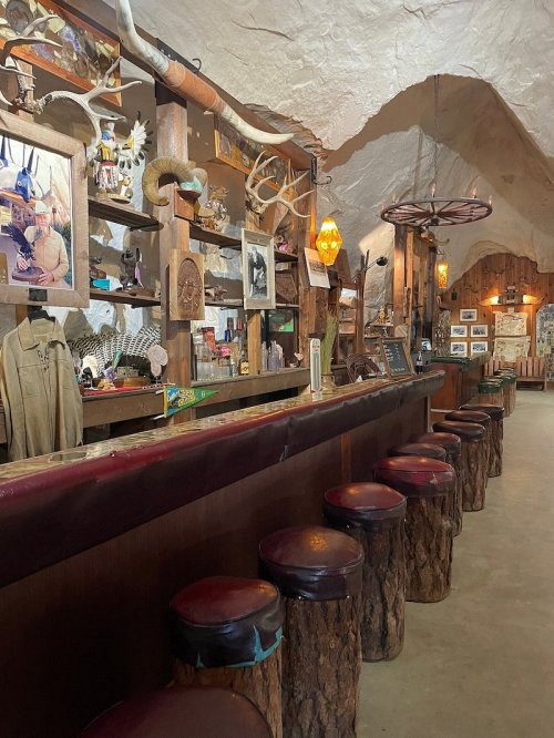 A rustic bar with wooden stools, antler decor, and a warm, inviting atmosphere in a cave-like setting.