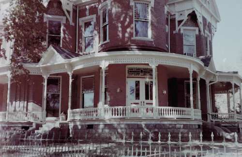Victorian-style brick house with a large porch, intricate details, and a decorative fence in front.