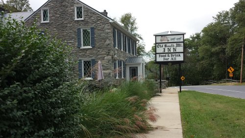 A stone building with a sign reading "Old South Mill Inn" and greenery along the sidewalk near a road.