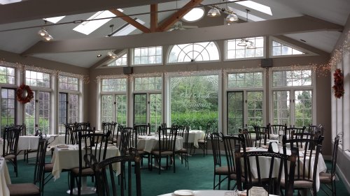 A bright dining room with tables set for guests, large windows, and festive decorations. Greenery visible outside.