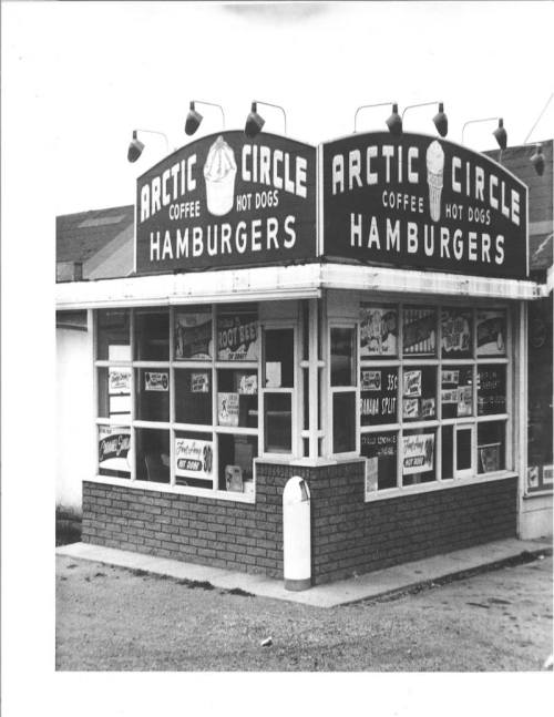 Black and white image of an Arctic Circle restaurant featuring signs for hamburgers, coffee, and hot dogs.