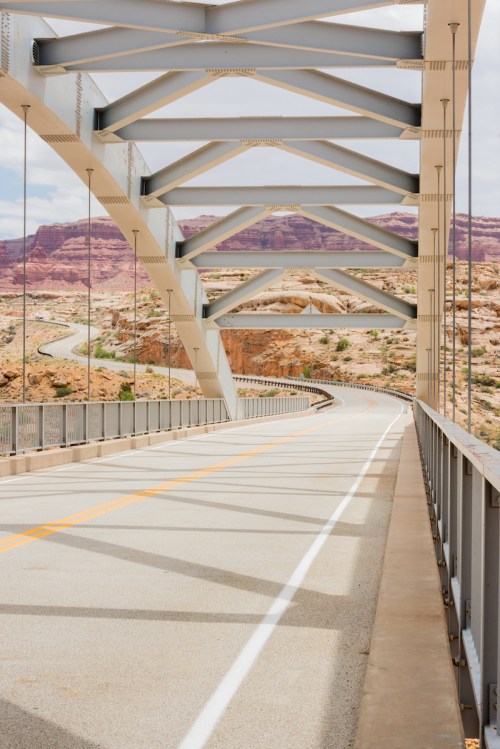 A view of a modern bridge with metal arches, leading to a winding road and rocky landscape in the background.