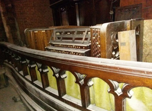 An old, dusty organ sits behind a wooden railing in a dimly lit, abandoned space.