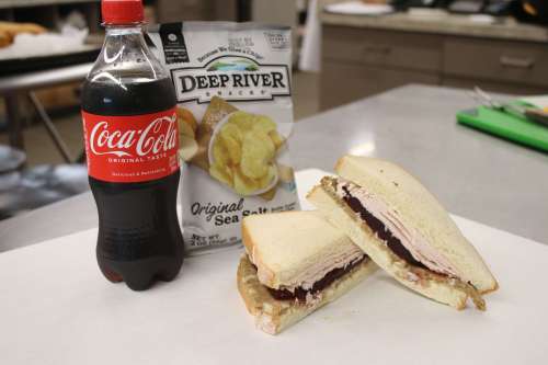 A sandwich with turkey and jelly, alongside a bottle of Coca-Cola and a bag of Deep River potato chips.