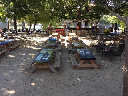 Outdoor seating area with wooden picnic tables and chairs under trees, featuring colorful table designs.