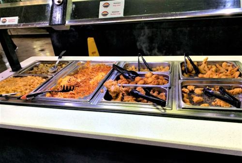 A buffet display featuring various dishes, including pasta, fried chicken, and shrimp, with serving tongs.