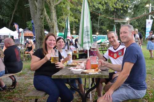 A group of four friends enjoying drinks and food at an outdoor festival, seated at a picnic table under trees.