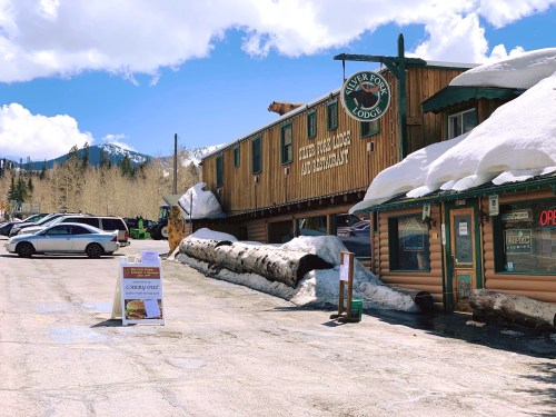 A rustic lodge with a sign reading "River Roll Lodge" and snow on the ground, surrounded by mountains and parked cars.