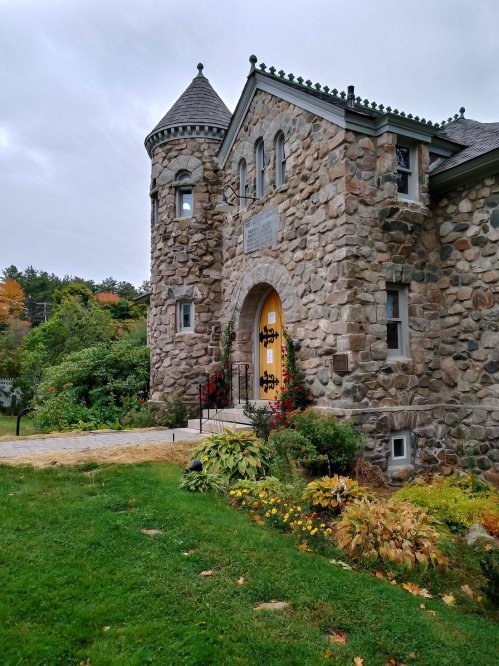 A stone building with a turret, surrounded by colorful gardens and trees, under a cloudy sky.