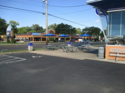 Outdoor seating area with blue umbrellas, near a road with shops in the background and clear blue sky.