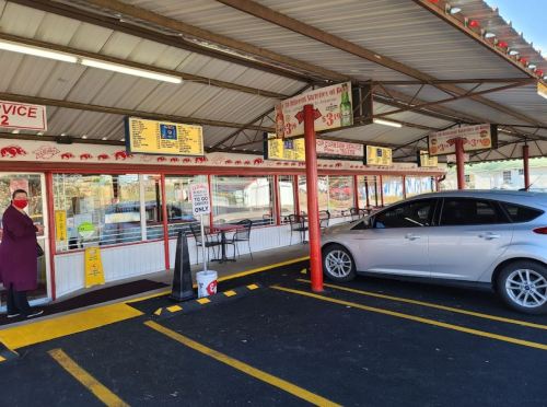 A drive-thru restaurant with a menu board, outdoor seating, and a silver car parked nearby. A person stands outside.