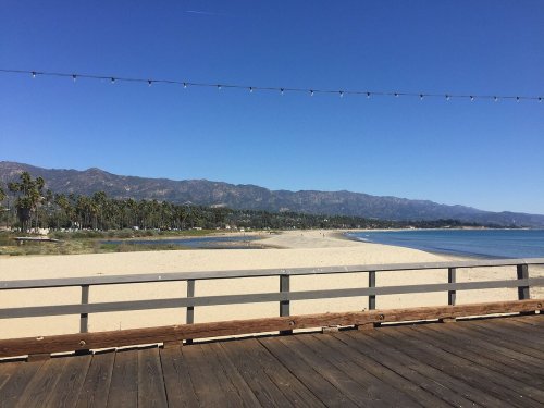 A sunny beach scene with a wooden pier, sandy shore, and mountains in the background under a clear blue sky.
