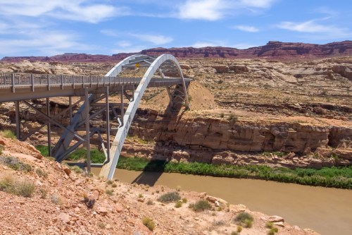 A modern arch bridge spans a river, surrounded by rocky cliffs and desert landscape under a blue sky.