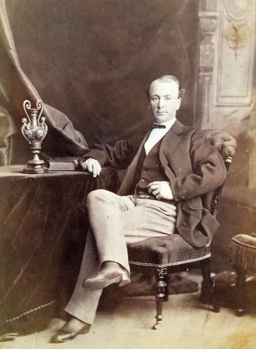 A seated man in 19th-century attire poses confidently in a studio, with a decorative vase beside him.