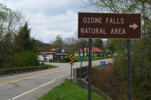 Sign for Ozone Falls Natural Area with a road and buildings in the background, under a cloudy sky.