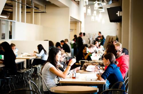 A busy cafeteria with people sitting at tables, eating and chatting in a bright, modern space.