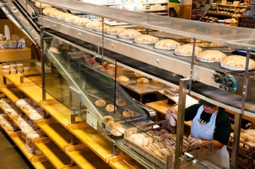 A bakery conveyor belt with freshly baked bread rolls being packaged, surrounded by shelves of baked goods.
