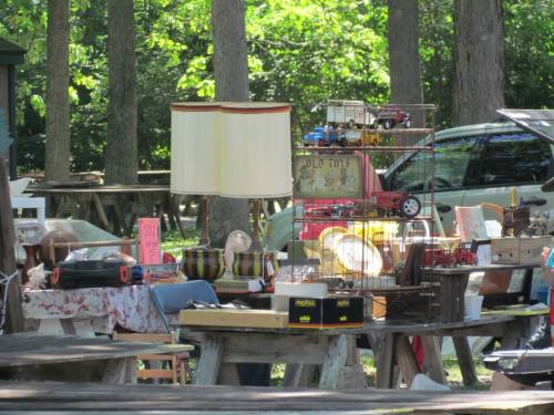 A bustling outdoor flea market with various items for sale, including a lamp, toys, and vintage decor on wooden tables.