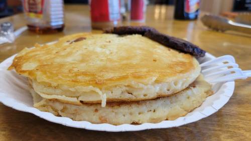 A plate with two fluffy pancakes and a piece of sausage, set on a wooden table.