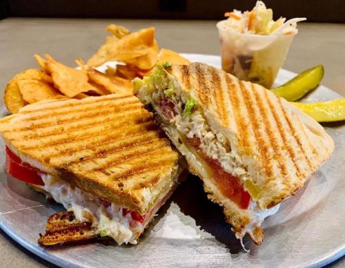 A toasted sandwich with chicken, tomato, and lettuce, served with chips, pickles, and a small cup of coleslaw.