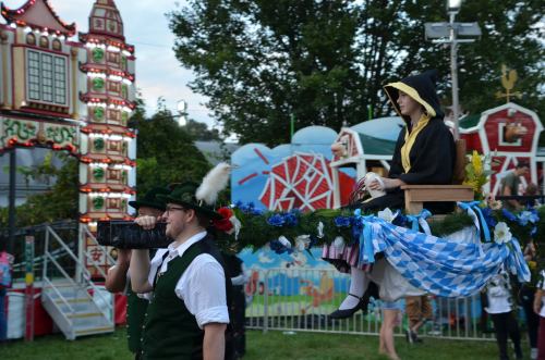 A person in a black and yellow outfit sits on a float, while another in traditional attire holds a camera at a fair.
