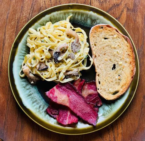 A plate of fettuccine with mushrooms, crispy bacon, and a slice of toasted bread on a wooden table.