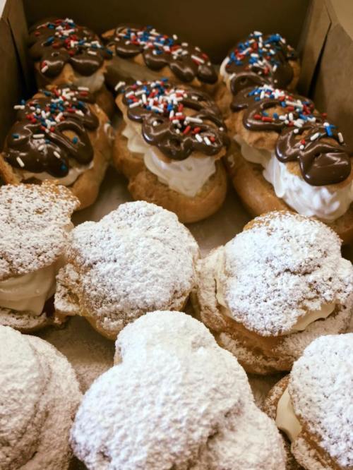 A box of assorted pastries: cream-filled eclairs topped with chocolate and sprinkles, and powdered sugar-covered cream puffs.