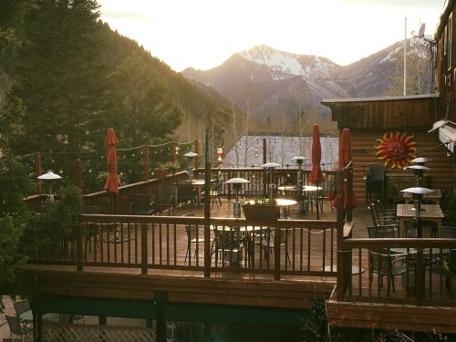 A wooden deck with tables and umbrellas, surrounded by mountains and trees, bathed in warm sunset light.
