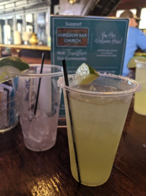 Two drinks on a wooden table, one with a lime garnish and salt rim, alongside a menu in a bar setting.